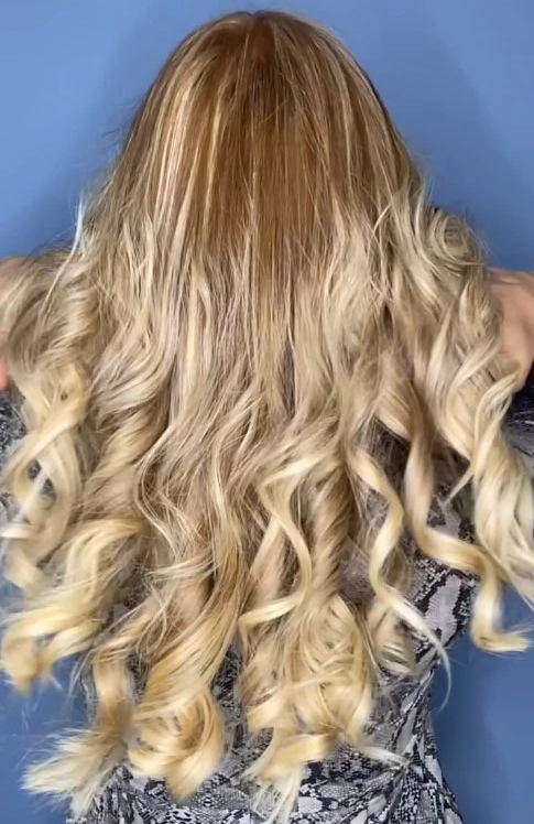 Capelli lunghi biondi con radici ambrate e onde morbide visti di spalle, maglia pitonata su sfondo azzurro